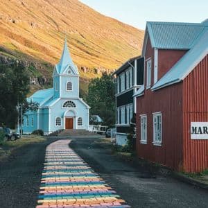 Un camino pintado de colores arcoíris en una carretera de asfalto conduce a una iglesia azul claro en la base de una montaña iluminada por el sol.