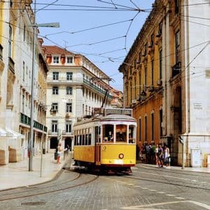 Un tram giallo percorre una strada lastricata che curva tra alti edifici classici europei sotto un cielo sereno.