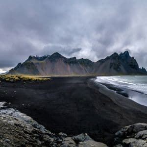 Una vista panorámica de una playa de arena negra con olas del océano llegando a la orilla frente a una escarpada cadena montañosa bajo un cielo nublado.