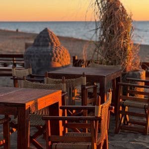 Tavoli e sedie di legno vuoti su una spiaggia sabbiosa al tramonto, con una grande testa di Buddha in pietra e l'oceano sullo sfondo.