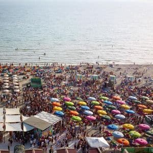 Una vista aerea di una festa in spiaggia affollata con persone sotto ombrelloni colorati e che nuotano nel mare calmo.