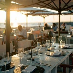 Un lungo tavolo da pranzo apparecchiato con bicchieri e lampade in un ristorante sulla spiaggia all'aperto al tramonto.