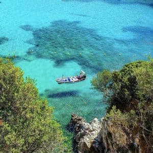 Vue en plongée d'un petit bateau flottant sur une eau turquoise cristalline, aperçu entre des arbres verts le long d'une côte rocheuse.