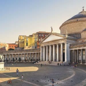 Una piazza pubblica assolata con un grande edificio a cupola e un colonnato classico, e persone e statue sparse tutt'intorno.