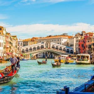 Des gondoles et d'autres bateaux naviguent sur un canal animé, avec un grand pont de pierre voûté et des bâtiments historiques colorés sous un ciel bleu.