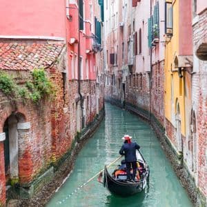 Un gondolier coiffé d'un chapeau rame sur une gondole le long d'un étroit canal entouré de bâtiments en briques colorées.