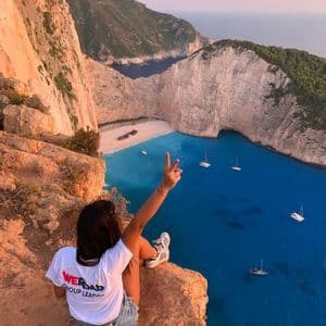 Un líder de grupo de WeRoad se sienta al borde de un acantilado con vistas a una cala con un naufragio en la playa y barcos en el agua turquesa.