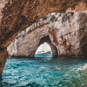 Velero blanco enmarcado por un arco de roca natural, visto desde el interior de una cueva marina sobre agua turquesa.