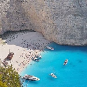Vista aérea de una playa concurrida con un naufragio oxidado al pie de altos acantilados blancos. Numerosas embarcaciones flotan en el agua turquesa.