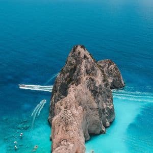 Vista aérea de una gran formación rocosa en una playa rodeada de agua turquesa. Varias embarcaciones navegan en el mar azul profundo cercano.