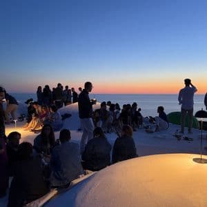 Un gruppo WeRoad socializza su un tetto bianco, ammirando il tramonto sul mare.