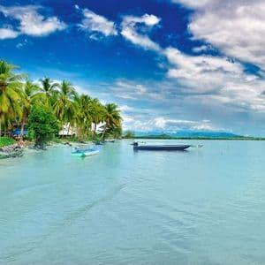 Una casa su palafitte colorata su una spiaggia sabbiosa fiancheggiata da palme, con piccole barche che galleggiano su acque calme e turchesi sotto un cielo blu.
