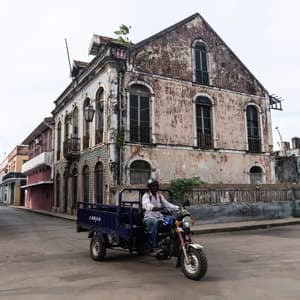 Un uomo percorre una strada a bordo di una moto a tre ruote blu, passando accanto a vecchi edifici in stile coloniale segnati dal tempo, sotto un cielo coperto.