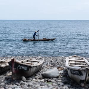 Un uomo su una barca di legno rema in mare, mentre altre tre barche sono parcheggiate su una spiaggia di ciottoli in primo piano.