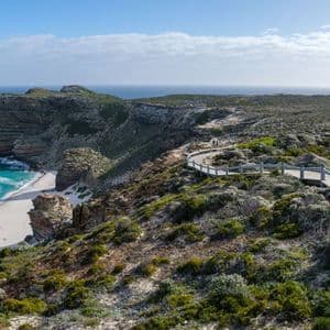 Une passerelle en bois serpente le long d'une falaise escarpée et verdoyante, surplombant une plage de sable blanc et un océan turquoise sous un ciel bleu.
