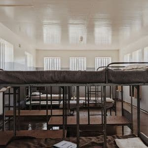 Intérieur d'un dortoir austère avec des rangées de lits superposés en métal, des couvertures en laine et des fenêtres à barreaux.