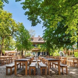 File di tavoli e panche di legno vuoti si trovano su un patio ghiaioso in una birreria all'aperto, ombreggiata da grandi alberi frondosi.