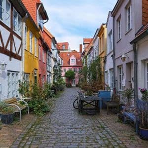 Un vicolo stretto di ciottoli fiancheggiato da case tradizionali e colorate, piante in vaso e mobili da esterno sotto un cielo parzialmente nuvoloso.