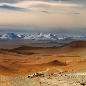 Una caravana de personas y ganado se mueve por un valle árido por un camino de tierra, con montañas nevadas al fondo.