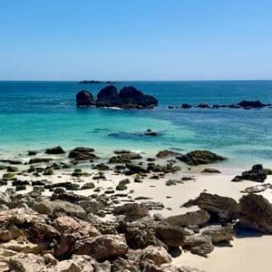 Una costa rocciosa degrada in una spiaggia di sabbia bianca, lambita da acque turchesi e blu, sotto un cielo sereno.