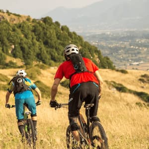 Due ciclisti percorrono in mountain bike un sentiero erboso in discesa, con una valle e montagne visibili in lontananza.