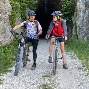 Due ciclisti con i caschi si fermano con le loro biciclette su un sentiero sterrato, sorridendosi davanti all'imbocco di una galleria buia.