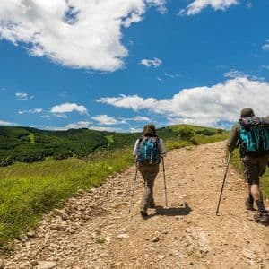 Due persone con zaini e bastoncini da trekking percorrono un sentiero sterrato su una collina erbosa, sotto un cielo azzurro con nuvole.