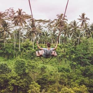 Une femme, vue de dos, se balance sur une grande balançoire très haut au-dessus d'une jungle luxuriante remplie de palmiers.