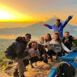 A WeRoad group trip smiling for a photo on a mountain summit during sunrise, with a volcanic mountain in the background.