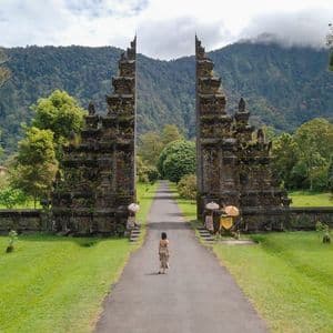 A woman seen from behind walks on a path towards an ornate split stone gate, with lush green trees and a mountain in the background.