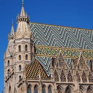 Gros plan sur la flèche de pierre ornée et le toit de tuiles colorées aux motifs géométriques d'une cathédrale, sous un ciel bleu clair.