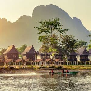 Una barca longtail con tre persone naviga su un fiume, costeggiando un villaggio di capanne di legno, con le montagne sullo sfondo durante un tramonto velato.