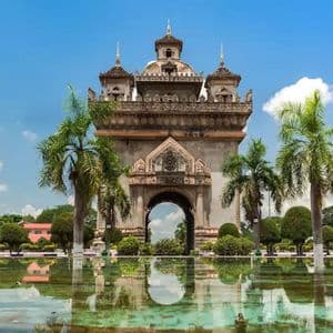Un arco in pietra finemente decorato, riflesso in una piscina, circondato da palme e giardini sotto un cielo azzurro.