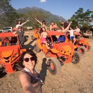 Un gruppo WeRoad si scatta un selfie mentre posa su e intorno a diversi dune buggy arancioni su una strada sterrata, con le montagne sullo sfondo.