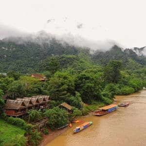 Palafitte in legno e barche longtail riposano sulla riva di un fiume fangoso ai piedi di una montagna coperta di nuvole.