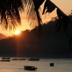 Barche navigano su un fiume durante un tramonto dorato, con il sole che scende dietro colline boscose e fronde di palma in silhouette in primo piano.