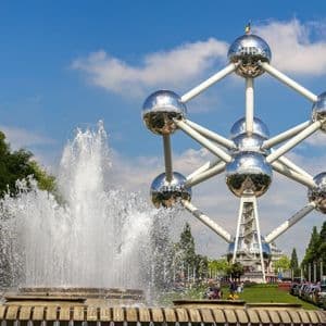 Le monument de l'Atomium se dresse derrière une grande fontaine d'eau dans un parc sous un ciel bleu avec des nuages épars.