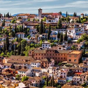Un denso pueblo en la ladera con edificios blancos, tejados de terracota y altos árboles verdes bajo un cielo parcialmente nublado.