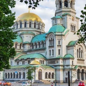 Eine prächtige weiße Kathedrale mit großen goldenen und kleineren türkisfarbenen Kuppeln, gesehen hinter den Blättern grüner Bäume.