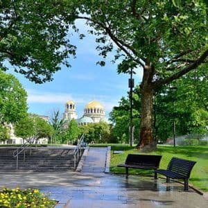 Ein Blick aus einem üppigen Stadtpark mit Steinwegen und Bänken, der eine ferne Kathedrale mit großen goldenen Kuppeln unter blauem Himmel umrahmt.