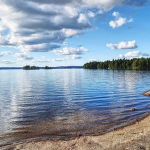 Ein breiter, ruhiger See mit felsigem Ufer unter einem teilweise bewölkten blauen Himmel, mit kieferbewachsenen Inseln in der Ferne.