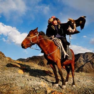 Una persona con vestimenta tradicional a caballo sostiene una gran águila en su brazo en una ladera rocosa bajo un cielo parcialmente nublado.