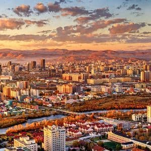 Una vista panorámica de una densa silueta urbana bañada por la luz dorada del atardecer, con montañas en el horizonte y un río abajo.