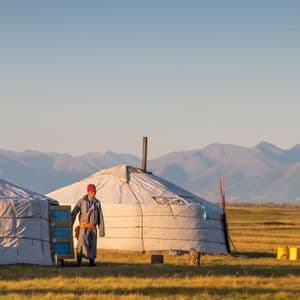 Una persona con ropa tradicional se encuentra junto a dos yurtas en un campo de hierba con una cordillera al fondo.