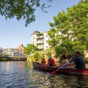 Eine vierköpfige Familie paddelt in einem roten Kanu auf einem Stadtfluss, vorbei an Bäumen und Gebäuden.