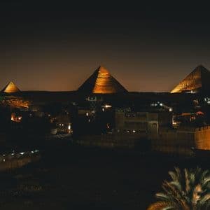 Vue nocturne de trois grandes pyramides illuminées de lumières dorées, s'élevant au-dessus d'un horizon urbain assombri.