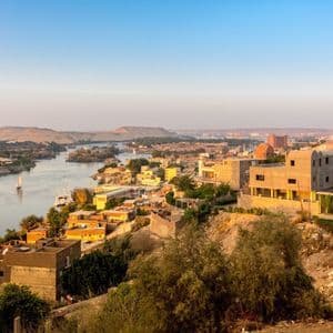 A high-angle view of a city with sand-colored buildings along a wide river with sailboats, with desert hills in the background.