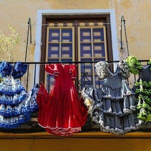 Cuatro vestidos de flamenca coloridos y con volantes colgando de un tendedero en un balcón frente a una ventana.