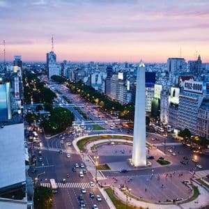 Una vista aerea di una città al tramonto, con un grande obelisco in una piazza circondata dal traffico su un ampio viale.