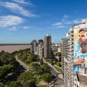 Un grande murale di Lionel Messi su un grattacielo, con vista su un paesaggio urbano verde e un ampio fiume sotto un cielo blu.